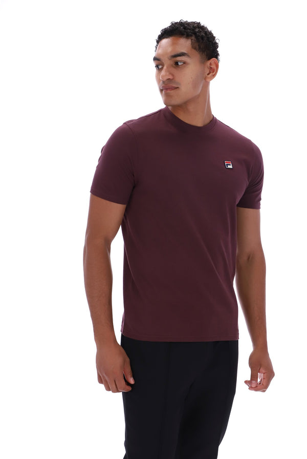 A man wearing a short-sleeve crewneck t-shirt in maroon with a small Fila logo on the left chest, paired with black pants, photographed against a solid white background.