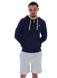 A male model wearing a navy blue zip-up hoodie with the FILA logo on the chest, paired with light grey shorts.
