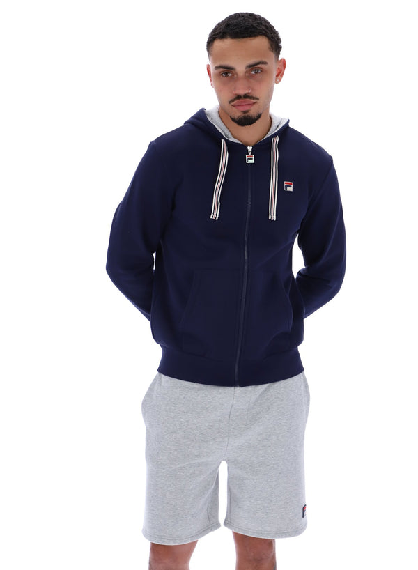 A male model wearing a navy blue zip-up hoodie with the FILA logo on the chest, paired with light grey shorts.