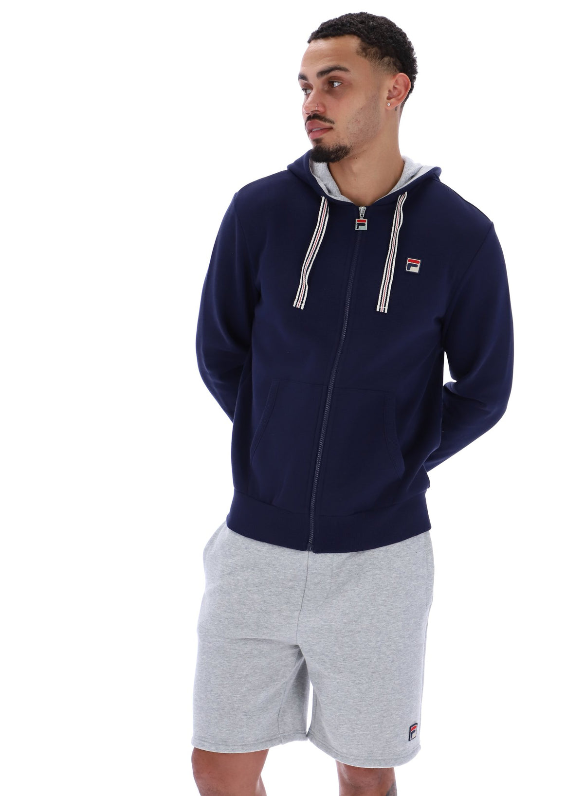 A male model wearing a navy blue zip-up hoodie with the FILA logo on the chest, paired with light grey shorts.