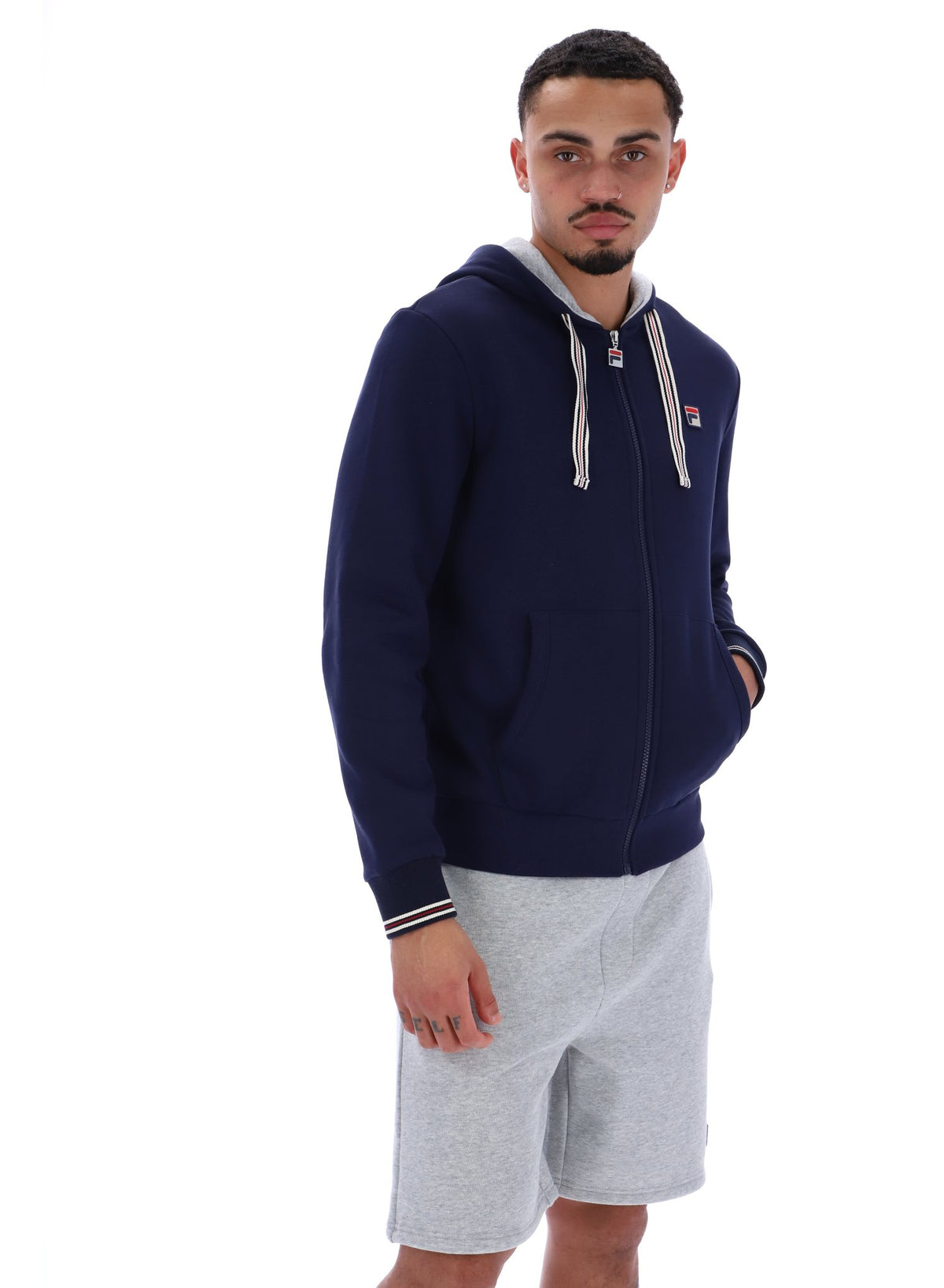 A male model wearing a navy blue zip-up hoodie with the FILA logo on the chest, paired with light grey shorts.