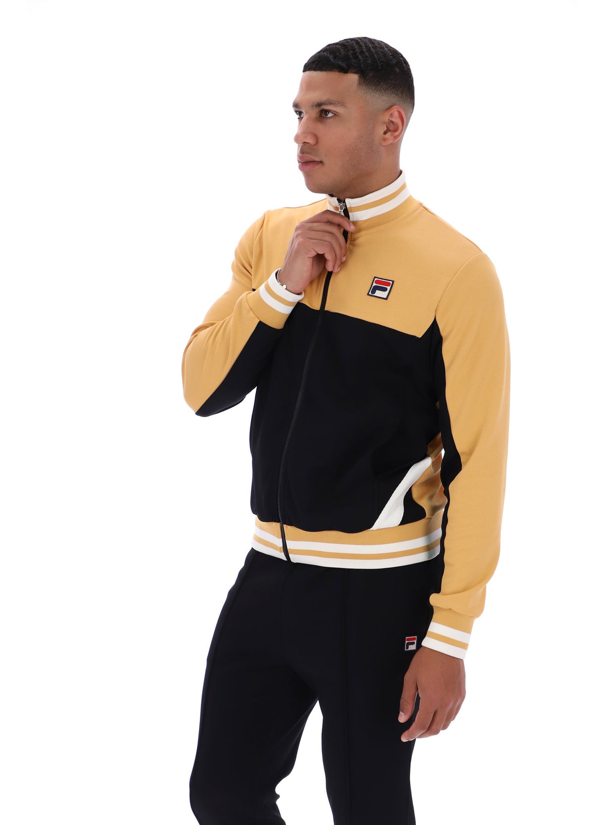 Man wearing a black and beige tracksuit with a FILA logo on the chest, paired with black FILA bottoms, against a white background