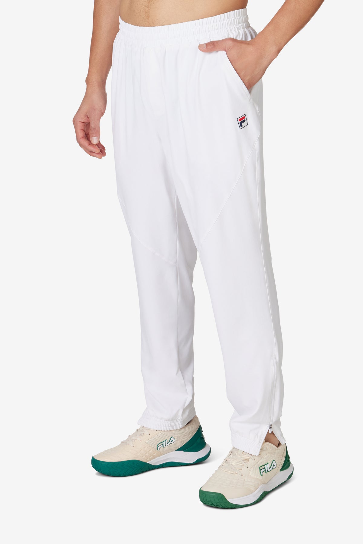 A person wearing white tennis court track pants with the FILA logo on the left leg and white sneakers.