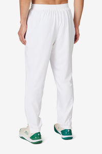 A person wearing white tennis court track pants with the FILA logo on the left leg and white sneakers.