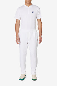 A person wearing white tennis court track pants with the FILA logo on the left leg and white sneakers.