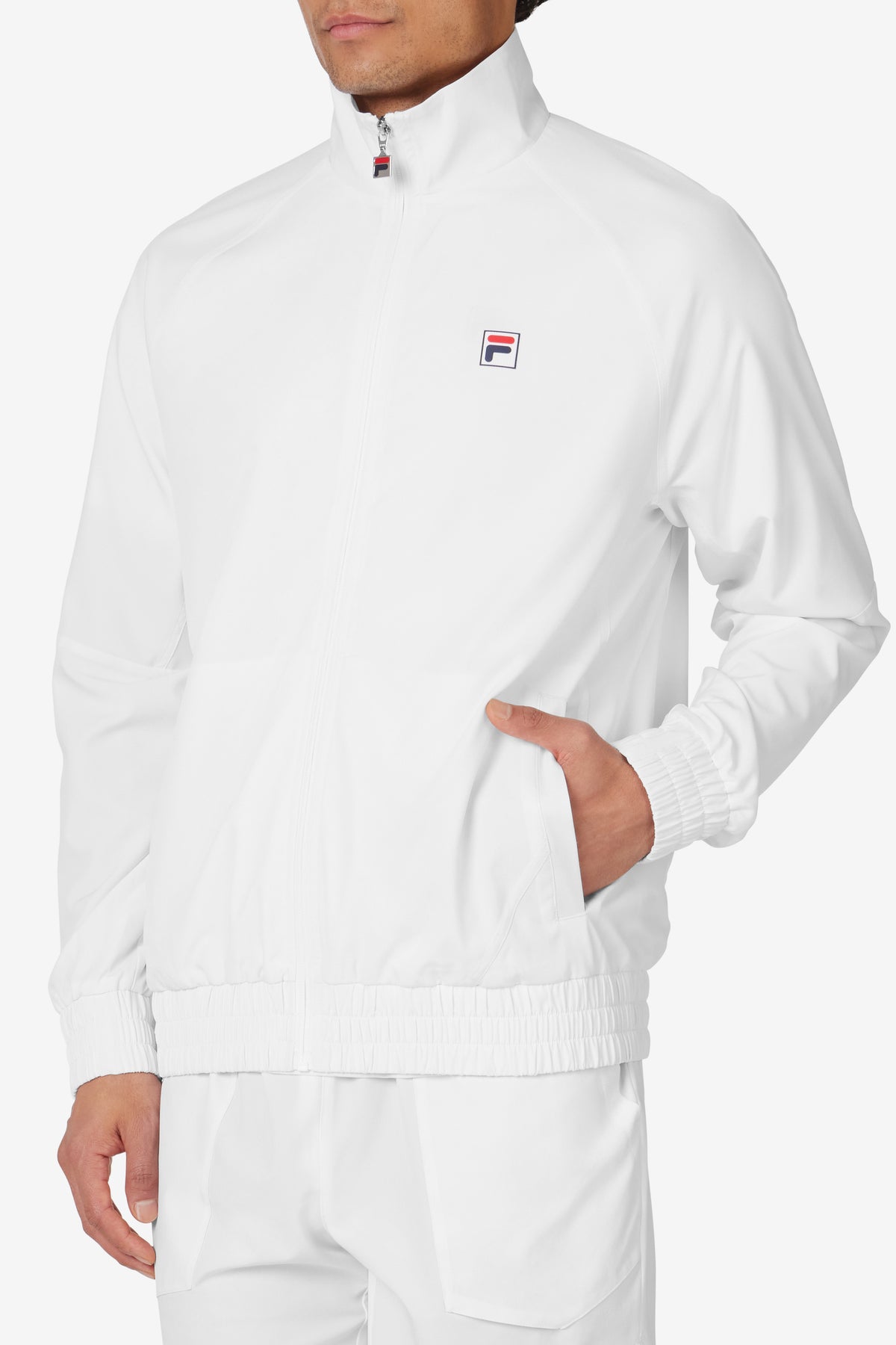 A man wearing a white FILA tennis court track jacket with a zipper, collar, and matching white pants.