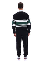 A man viewed from behind wearing a black knit sweater with green and white horizontal stripes across the shoulders and upper arms, paired with black sweatpants and white sneakers.