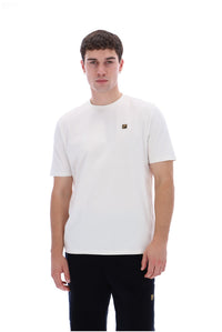 A man wearing a white crew-neck t-shirt with a small gold square logo on the left chest, paired with black pants, against a solid white background.