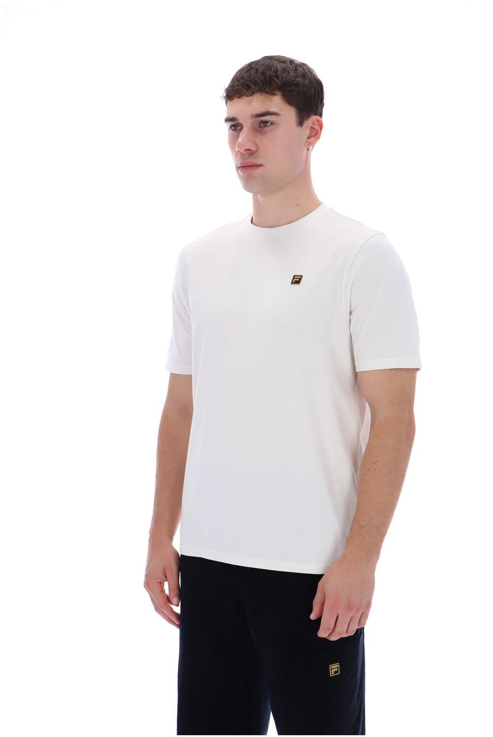A man wearing a white short-sleeve crewneck t-shirt with a small gold Fila logo on the left chest, paired with black pants, photographed against a solid white background.