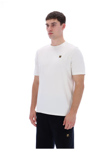 A man wearing a white short-sleeve crewneck t-shirt with a small gold Fila logo on the left chest, paired with black pants, photographed against a solid white background.