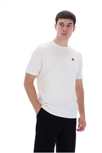 A man wearing a white short-sleeve crewneck t-shirt with a small gold square logo on the left chest, paired with black pants, against a solid white background.