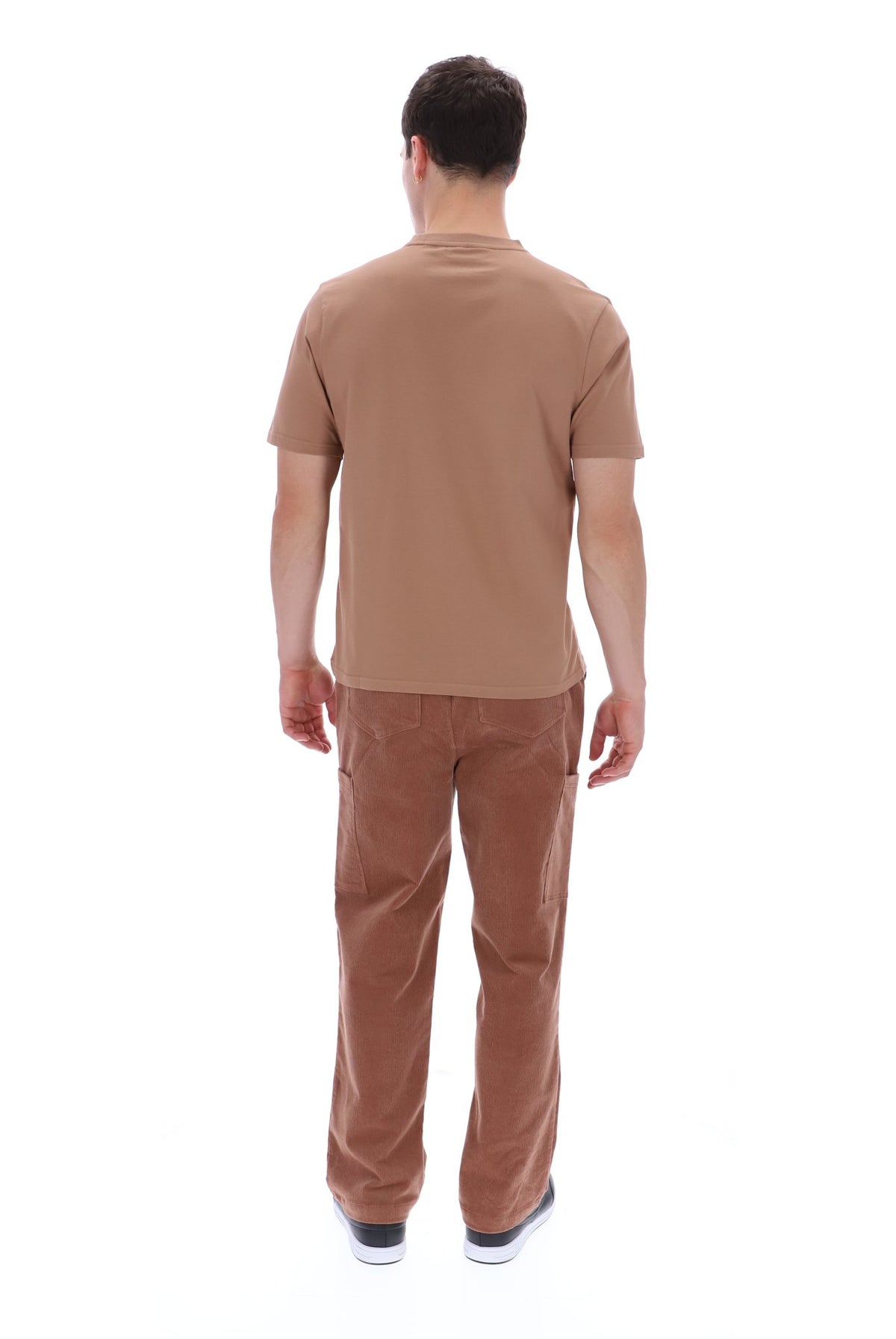 A man viewed from behind wearing a tan short-sleeve t-shirt and brown pants, standing against a plain white background.