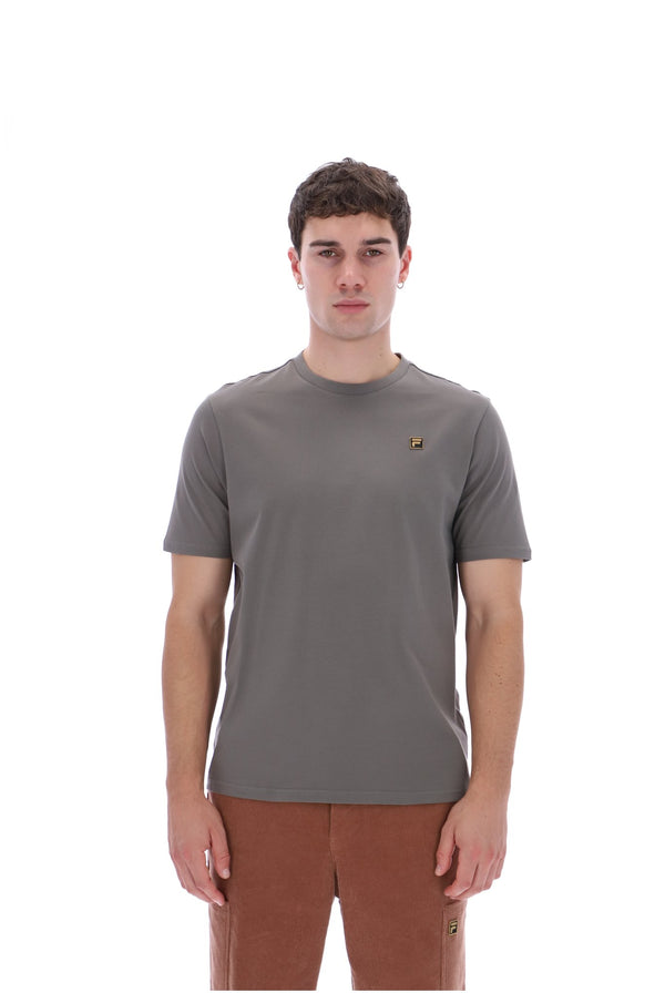 A man wearing a short-sleeve crewneck t-shirt in a muted gray-brown color, featuring a small embroidered logo on the left chest, paired with brown pants, against a solid white background.