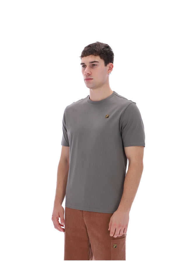 A man wearing a gray short-sleeve crewneck t-shirt with a small embroidered logo on the left chest, paired with brown pants, photographed against a solid white background.