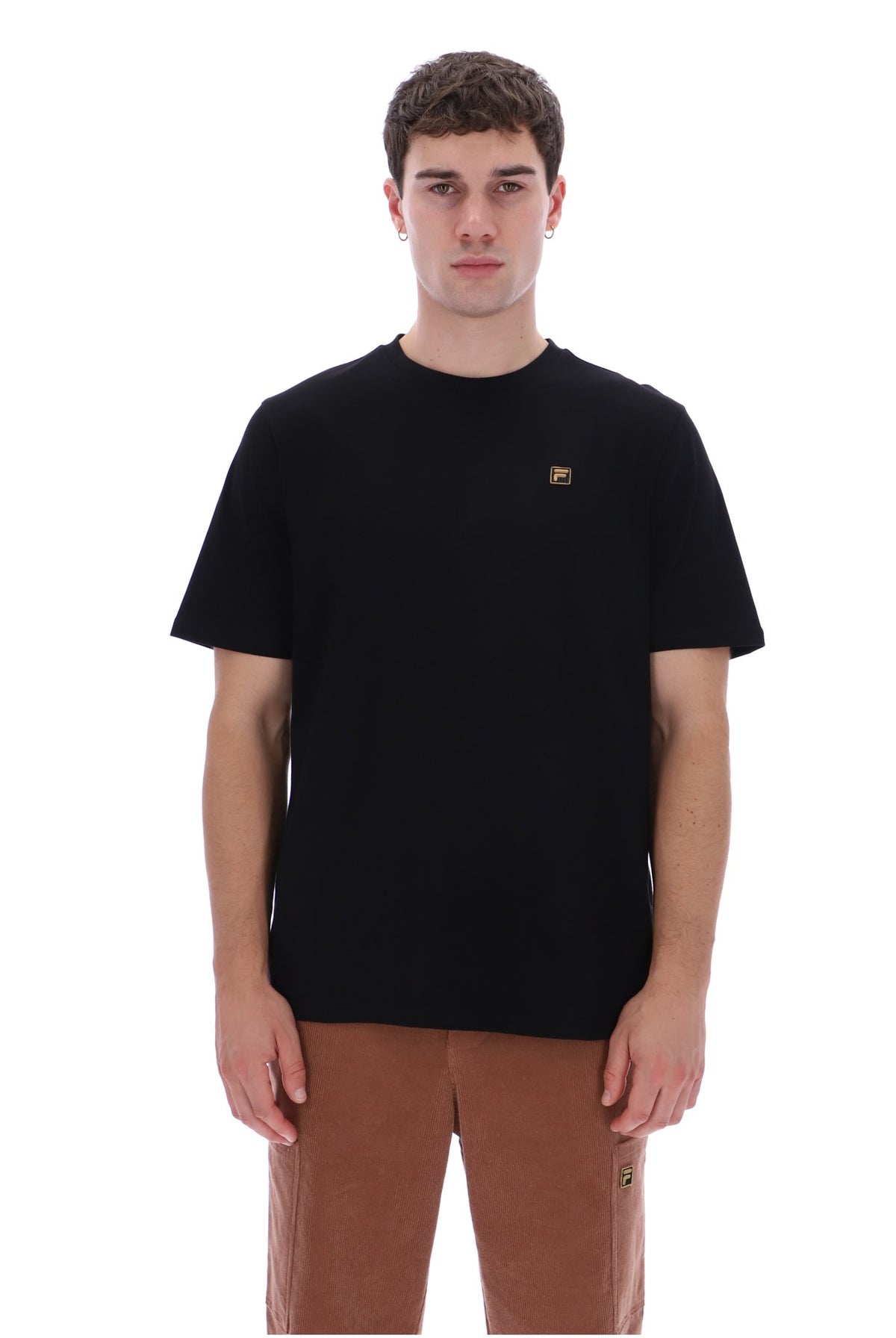 A man wearing a black short-sleeve crewneck t-shirt with a small gold logo on the left chest, paired with brown pants, photographed against a solid white background.