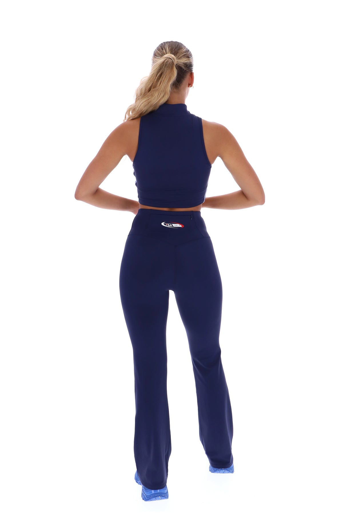 A woman wearing a navy blue zip-up crop top, paired with matching leggings that feature a FILA x TALA logo branded on the back.