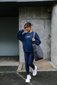 A person wearing high-waisted navy blue leggings with the FILA x TALA brand logo on the upper left thigh. They are also wearing a matching navy long-sleeve top and white sneakers, carrying a sports bag, and standing against an urban backdrop.