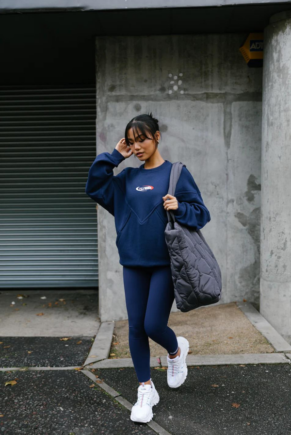 A person wearing high-waisted navy blue leggings with the FILA x TALA brand logo on the upper left thigh. They are also wearing a matching navy long-sleeve top and white sneakers, carrying a sports bag, and standing against an urban backdrop.